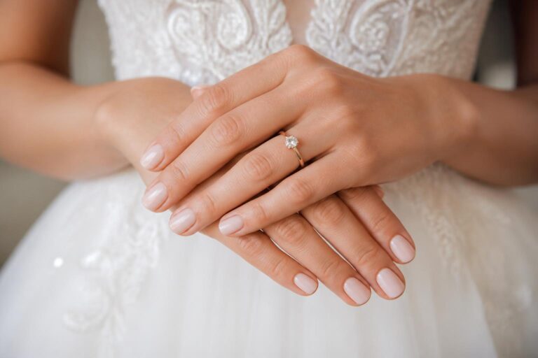 Short wedding nails with glossy nude pink polish and thin French tips, photographed close-up with an engagement ring